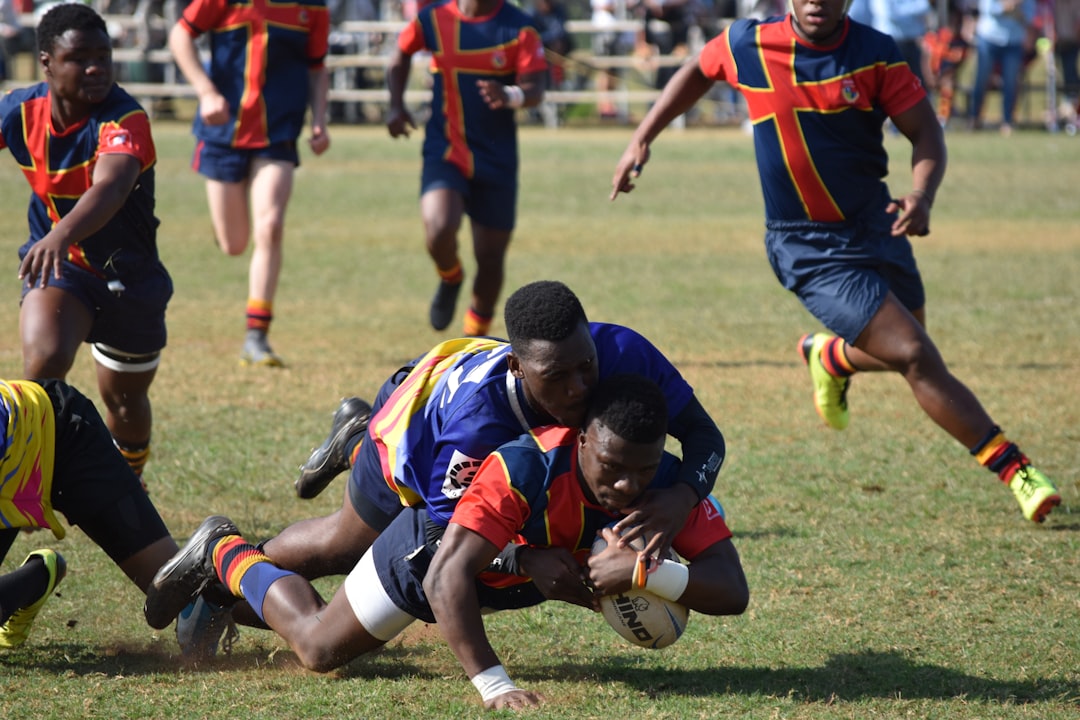 A young man (blue and yellow shirts) tackles his opponent (blue and red shirts) to the ground during a club rugby match in South Africa while the youngster clutches the ball and refuses to let go.